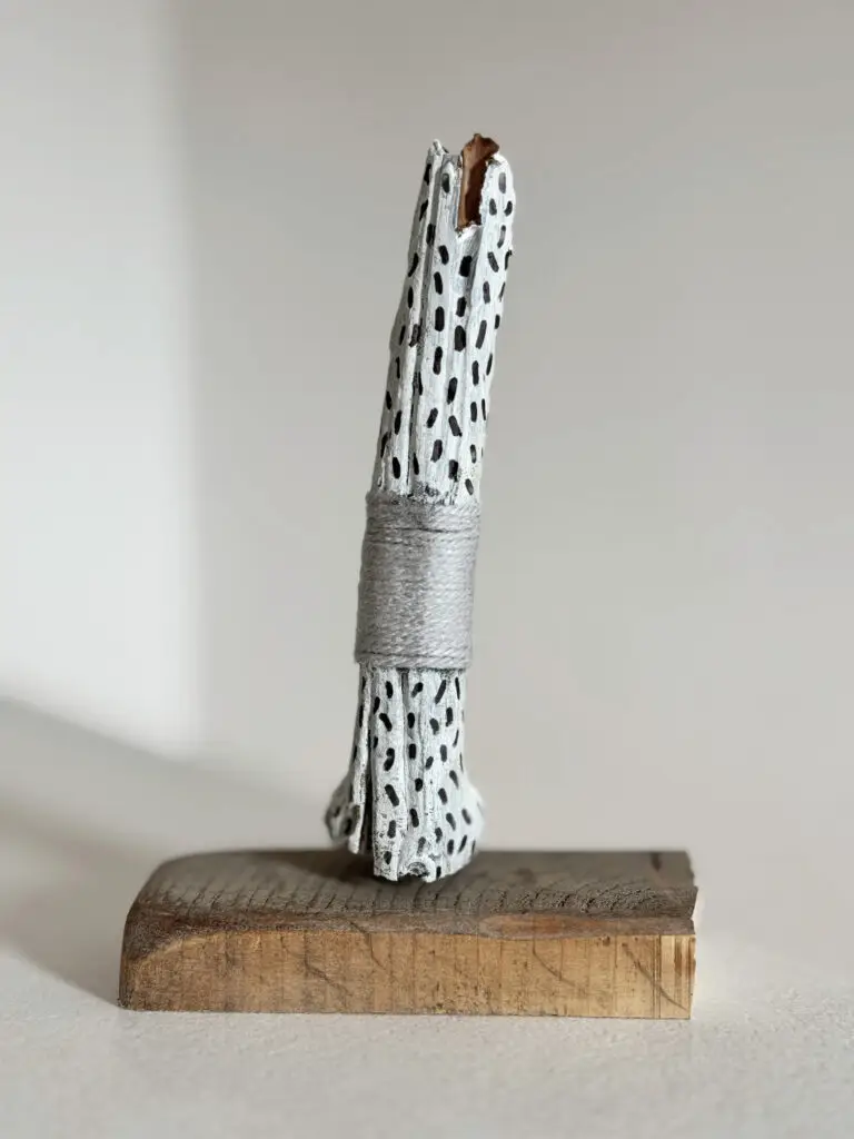 Bemalter Ast mit schwarzen Strichen und grauer Garnumwicklung auf Holzsockel